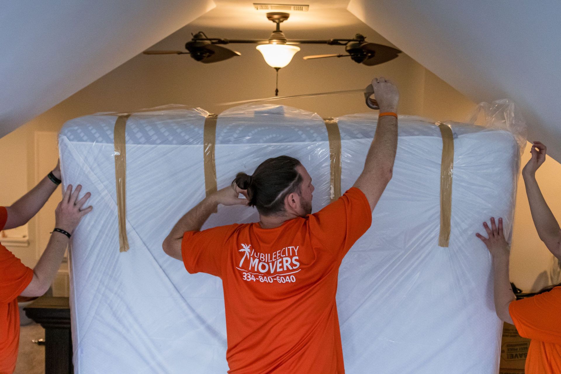 A man in an orange shirt is lifting a mattress in a room.