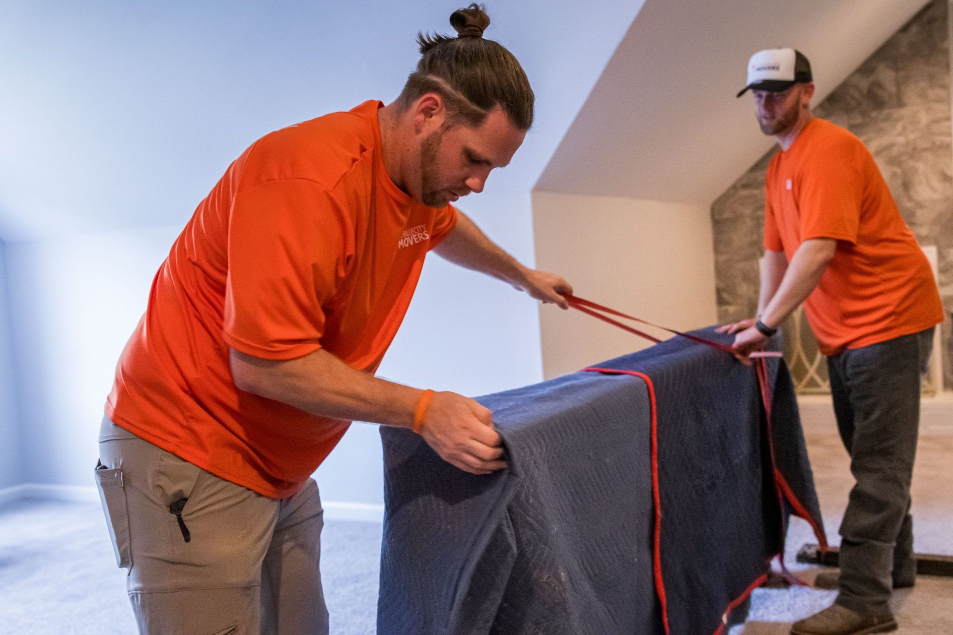 Two men in orange shirts are wrapping a piano in a blue blanket.