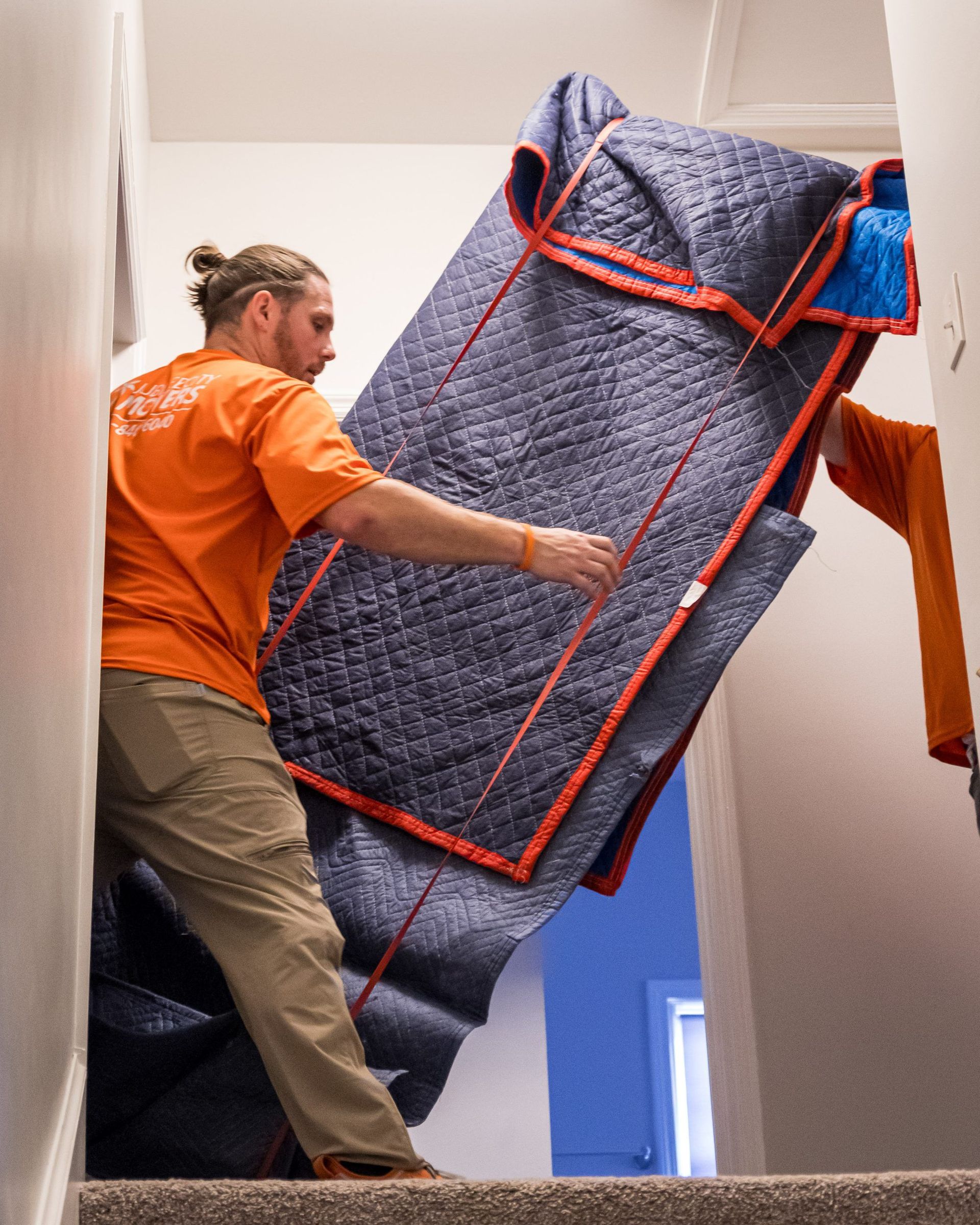 A man in an orange shirt is carrying a mattress up the stairs