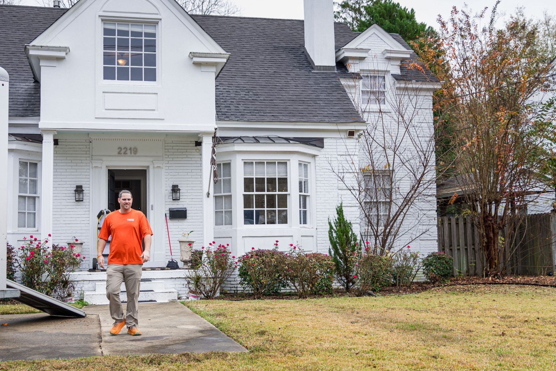 A man in an orange shirt is standing in front of a white house.