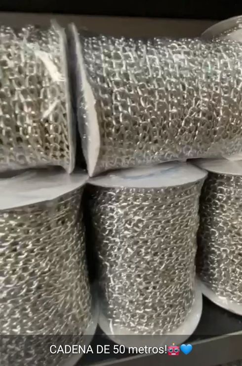 Rollos de malla metálica plateada apilados en un almacén, etiquetados como “cadena de 50 metros”.