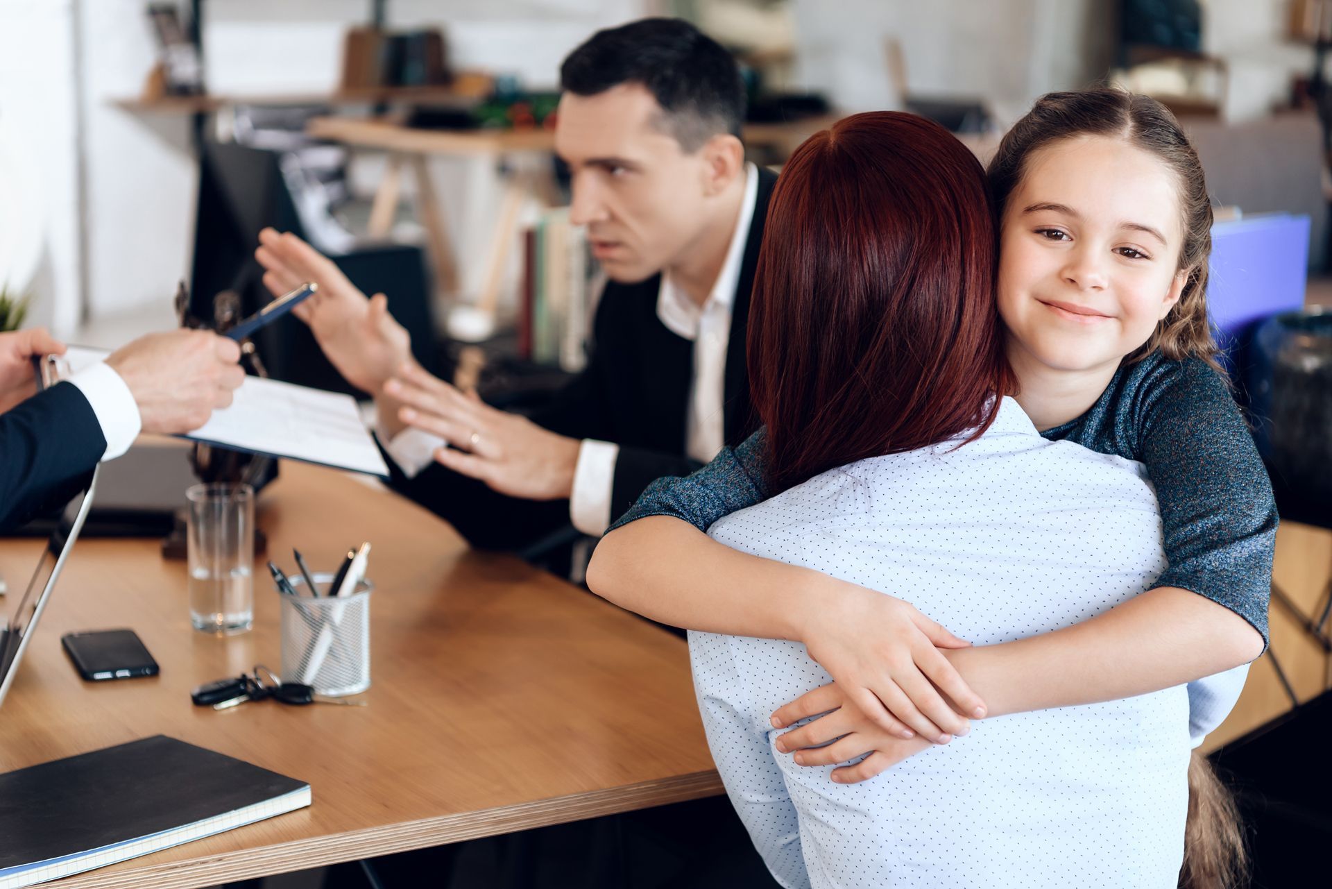Girl hugging a woman in an office; two men are at a table appearing displeased