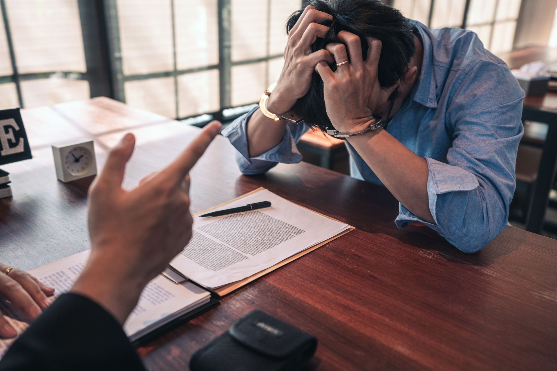 Man stressed, head in hands, being pointed at across a wooden desk, documents present
