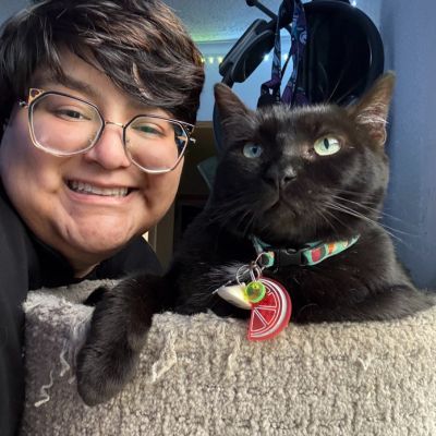 Person with glasses smiles next to a black cat on a cat tree; both look at the camera.