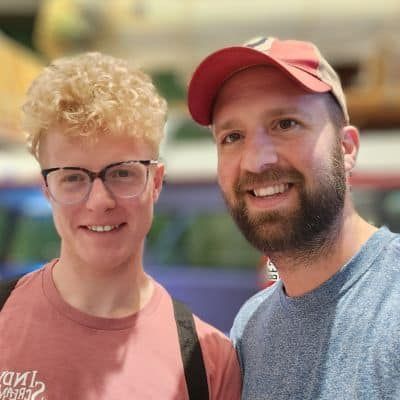 Two smiling people: one with curly blond hair and glasses, and one with a beard and a red cap.