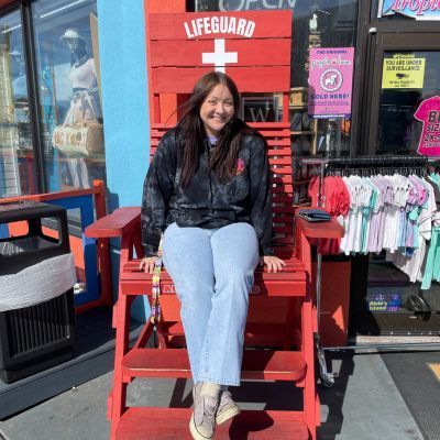 Woman sitting in a large red lifeguard chair, smiling, outside a shop.