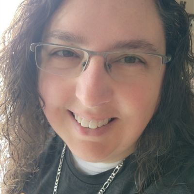 Person with curly brown hair, glasses, and a smiling expression. Wearing a silver chain, close-up shot.