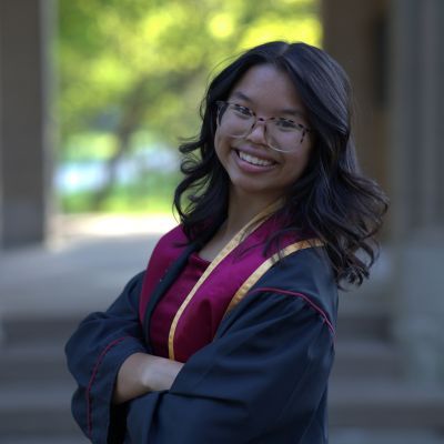 Woman in graduation attire, smiling with arms crossed, in a stone walkway. Burgundy sash, glasses, sunny background.