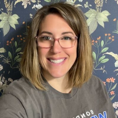 Woman with glasses, smiling, in front of floral wallpaper.