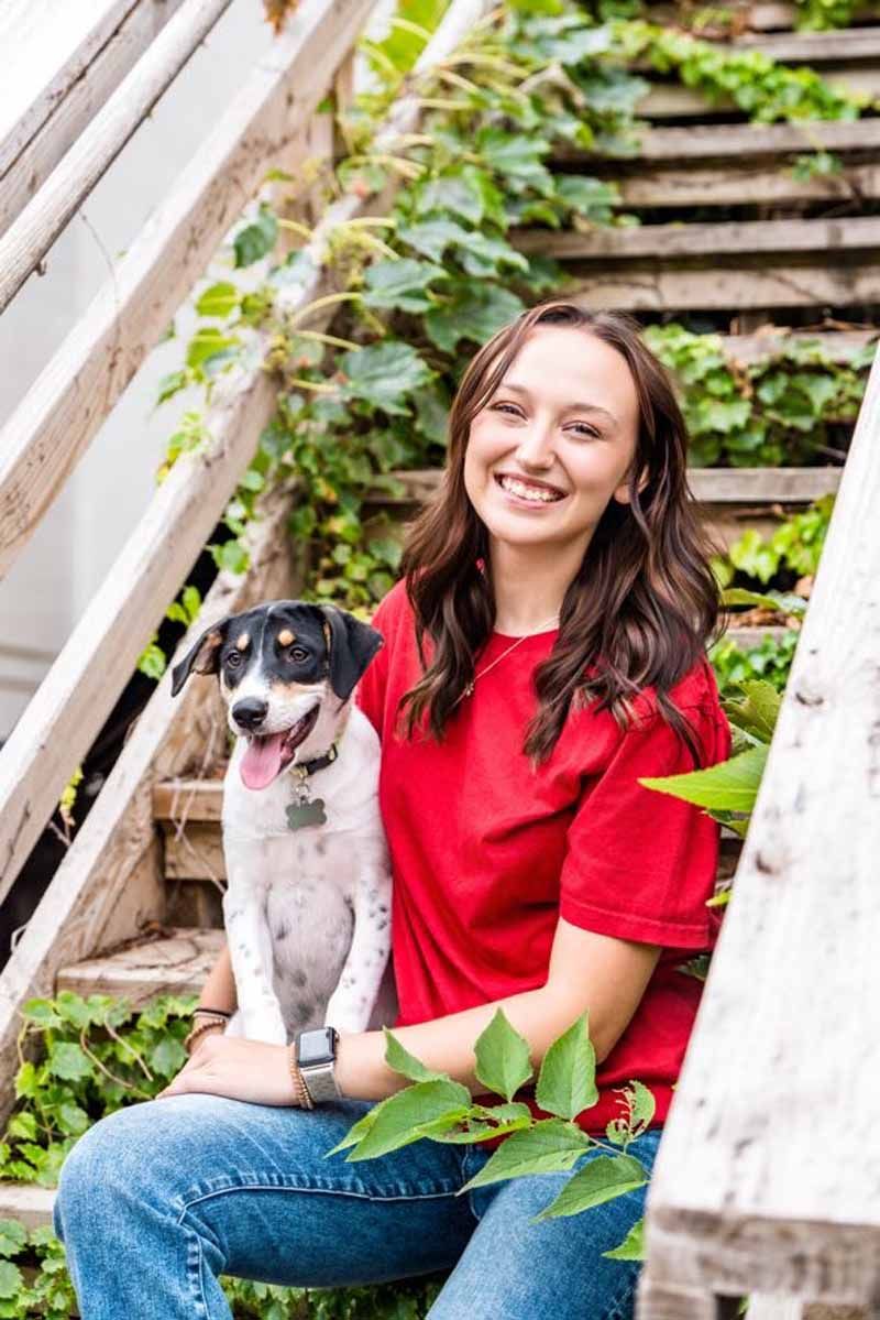Woman in red shirt and jeans sits on wooden stairs with a dog. Lush greenery surrounds them.