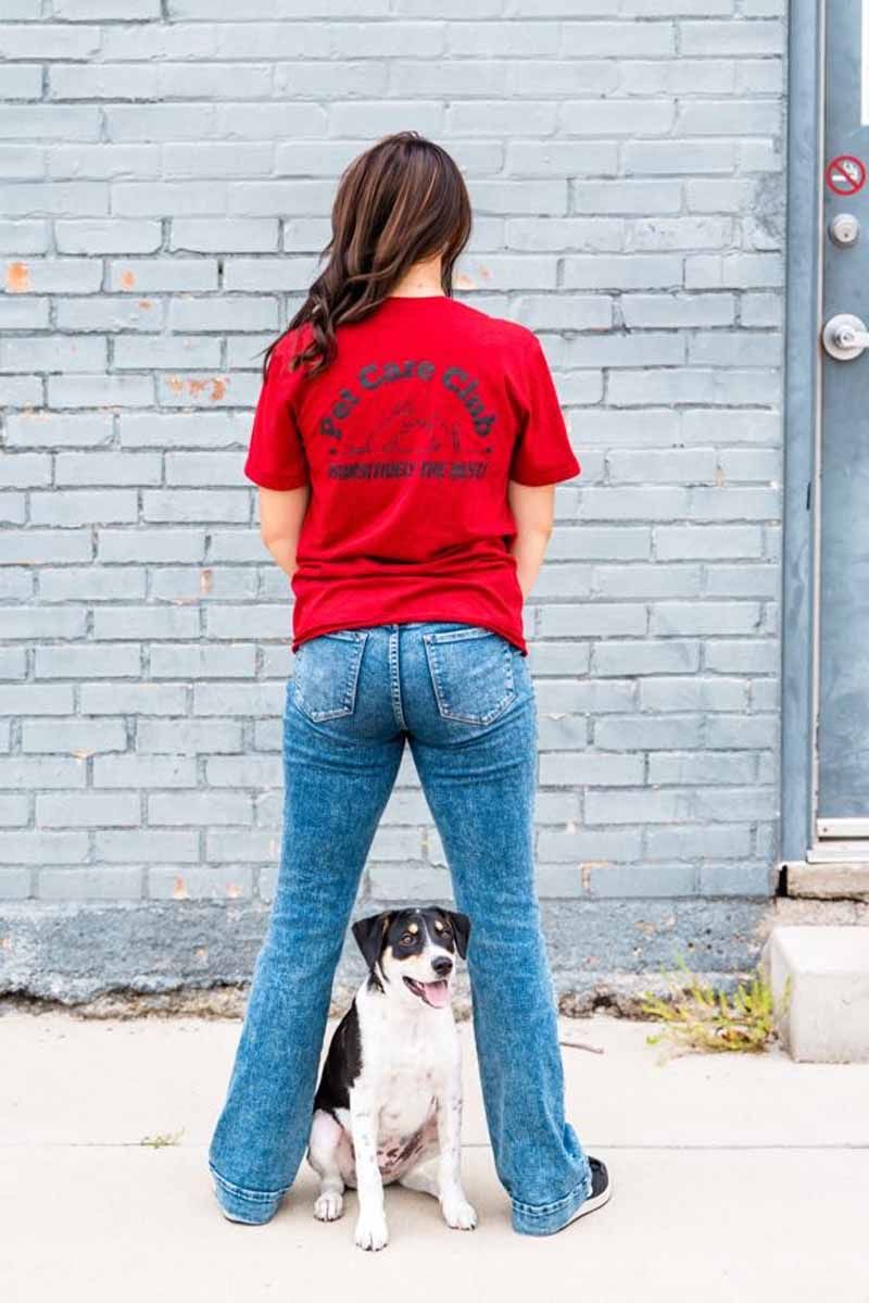 Woman in red shirt and jeans stands with legs spread, dog sits between her legs in front of a brick wall.