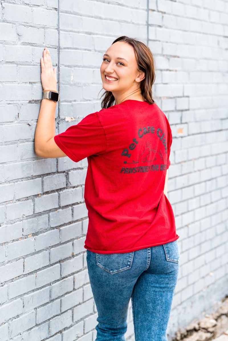 Woman in red t-shirt and jeans smiles, leaning against a gray brick wall, arm outstretched.