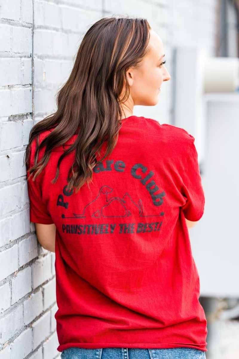 Woman leaning against a white brick wall, wearing a red t-shirt with black text on the back.