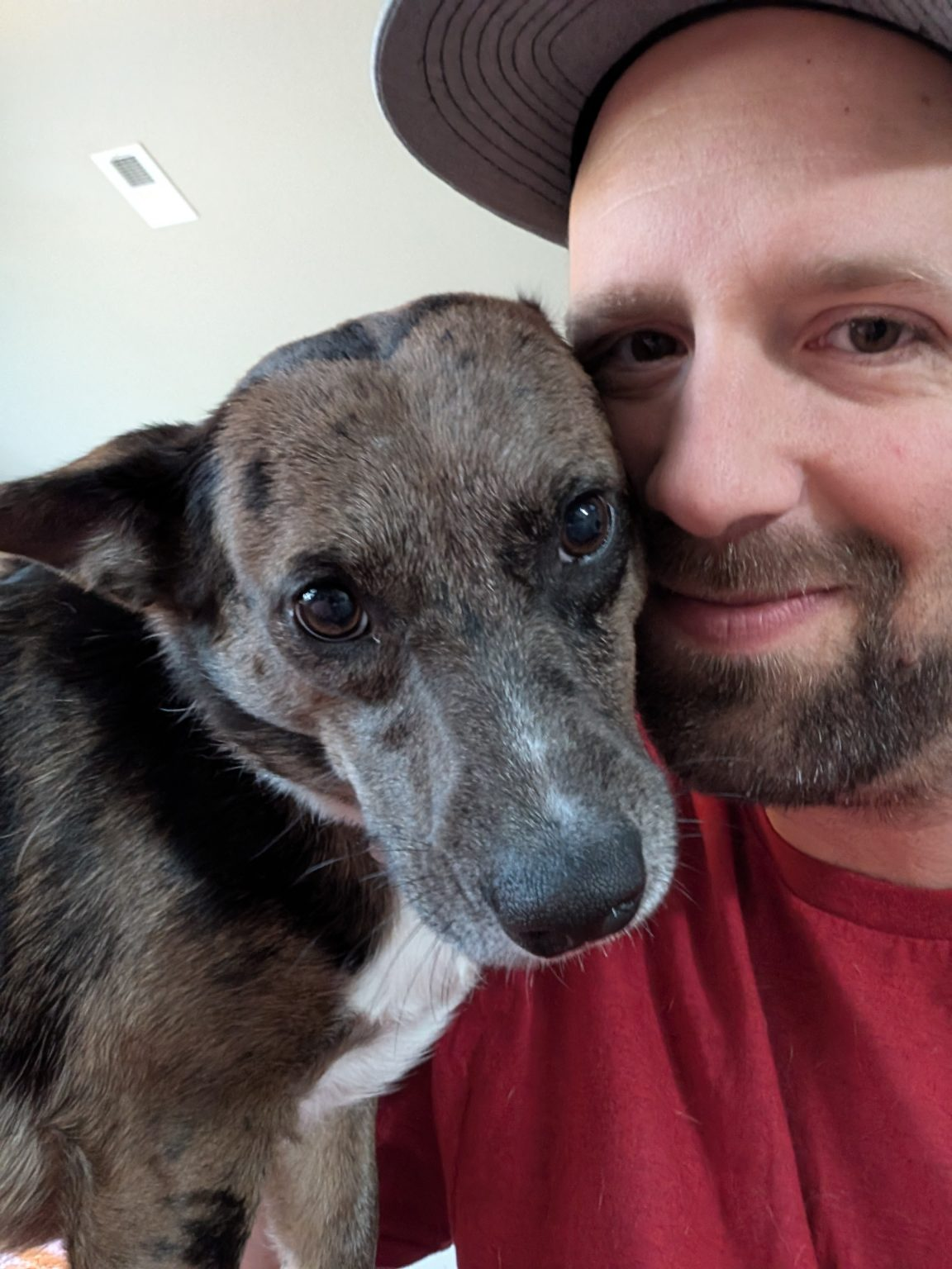 Man in red shirt and dog with mottled fur, smiling and close together.