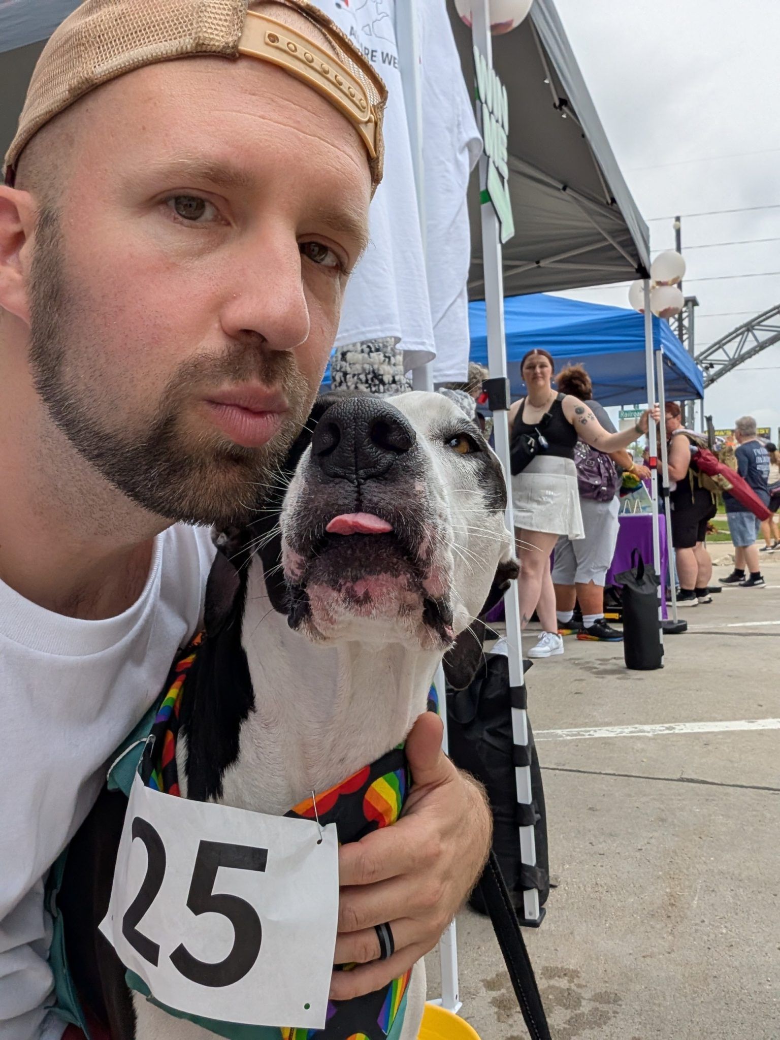 Man with dog sticking tongue out, wearing rainbow bib with number 25 at an outdoor event.
