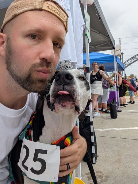 Man with dog sticking tongue out, wearing rainbow bib with number 25 at an outdoor event.