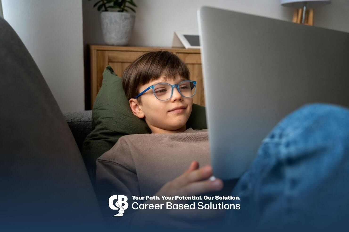 Autistic boy wearing glasses is sitting comfortably on a couch while looking at a laptop screen.