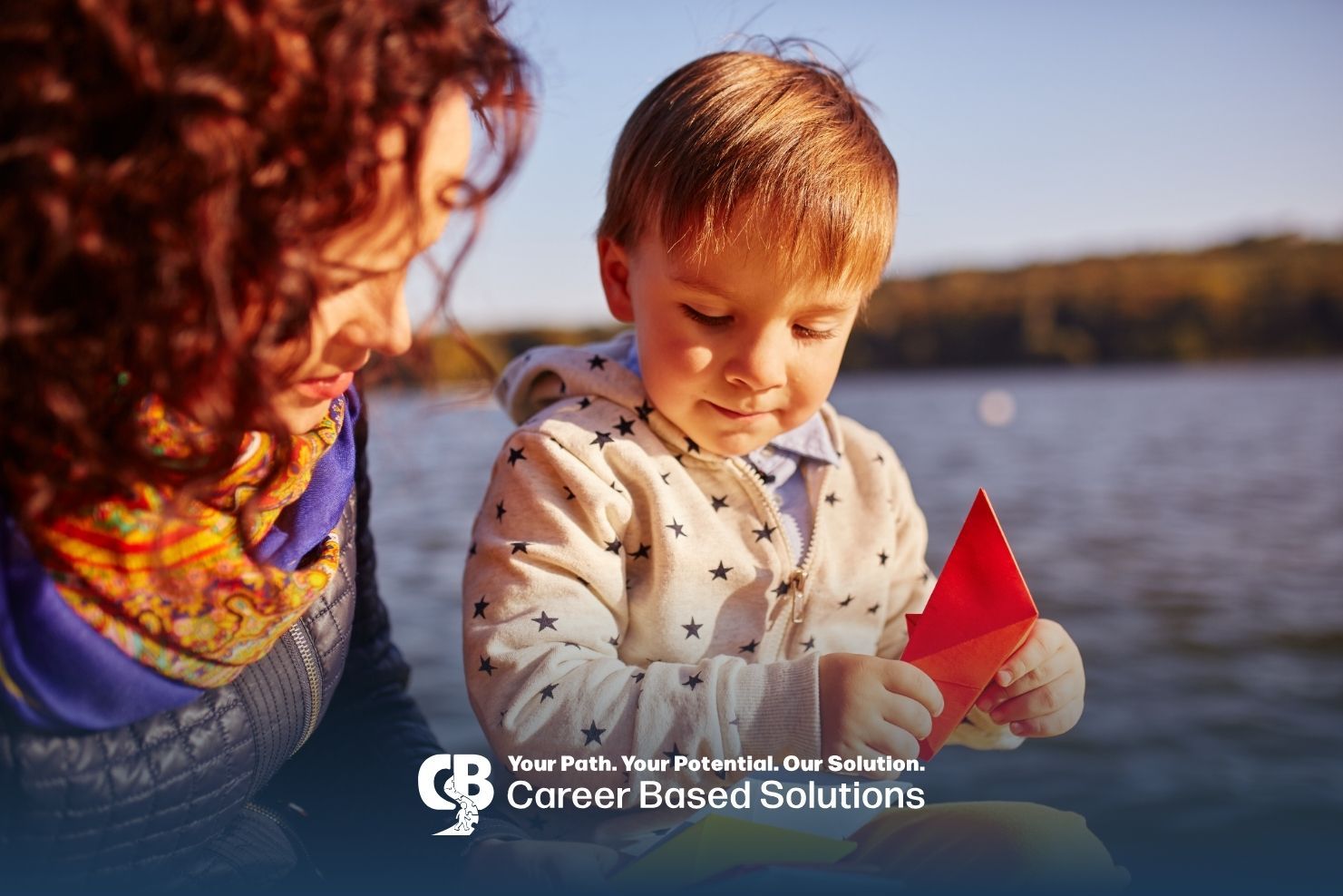 Therapist & autistic child fold colorful paper boats together beside a lake while enjoying summer.