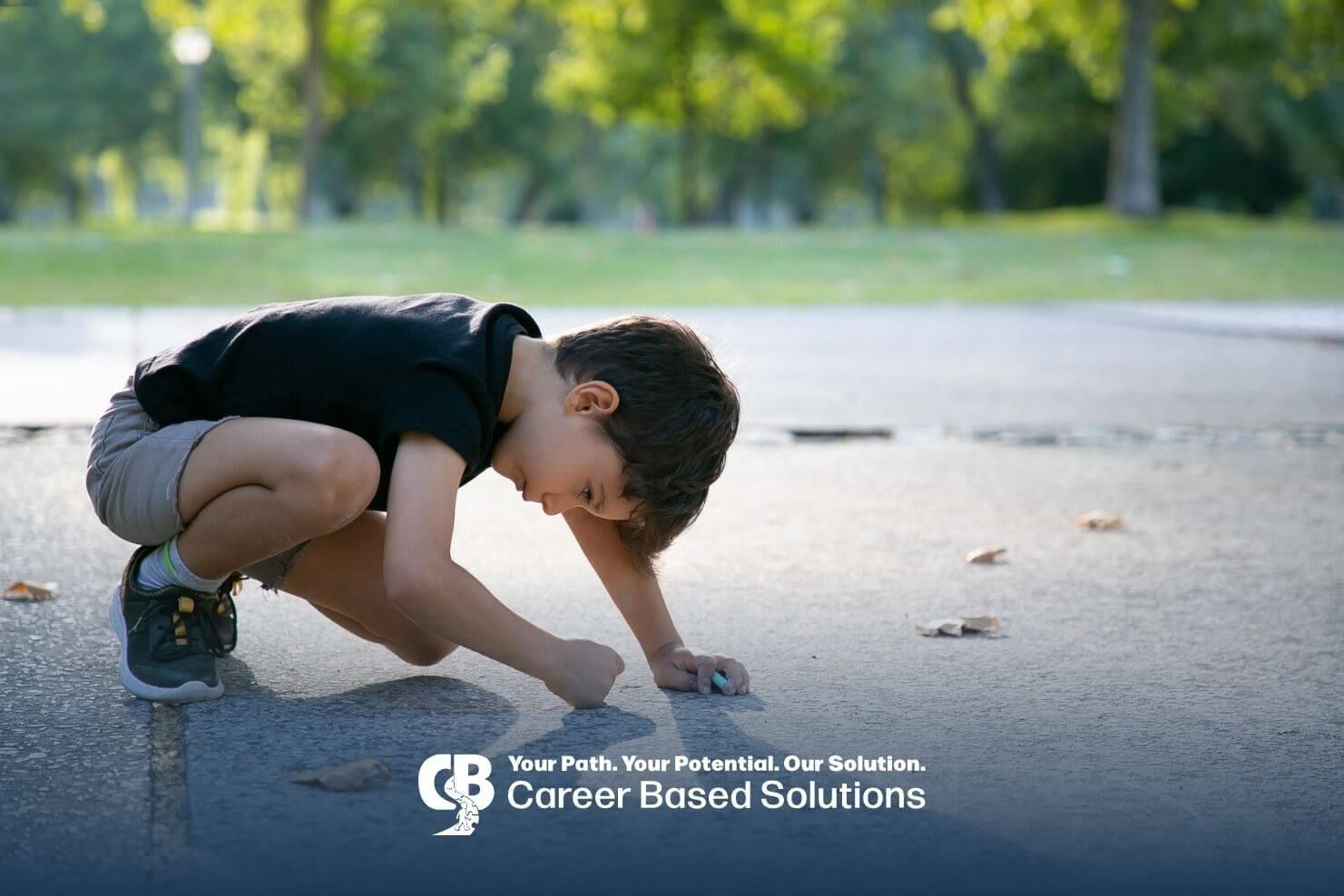 A young autistic boy crouching on a pavement drawing with chalk while focused on the ground.