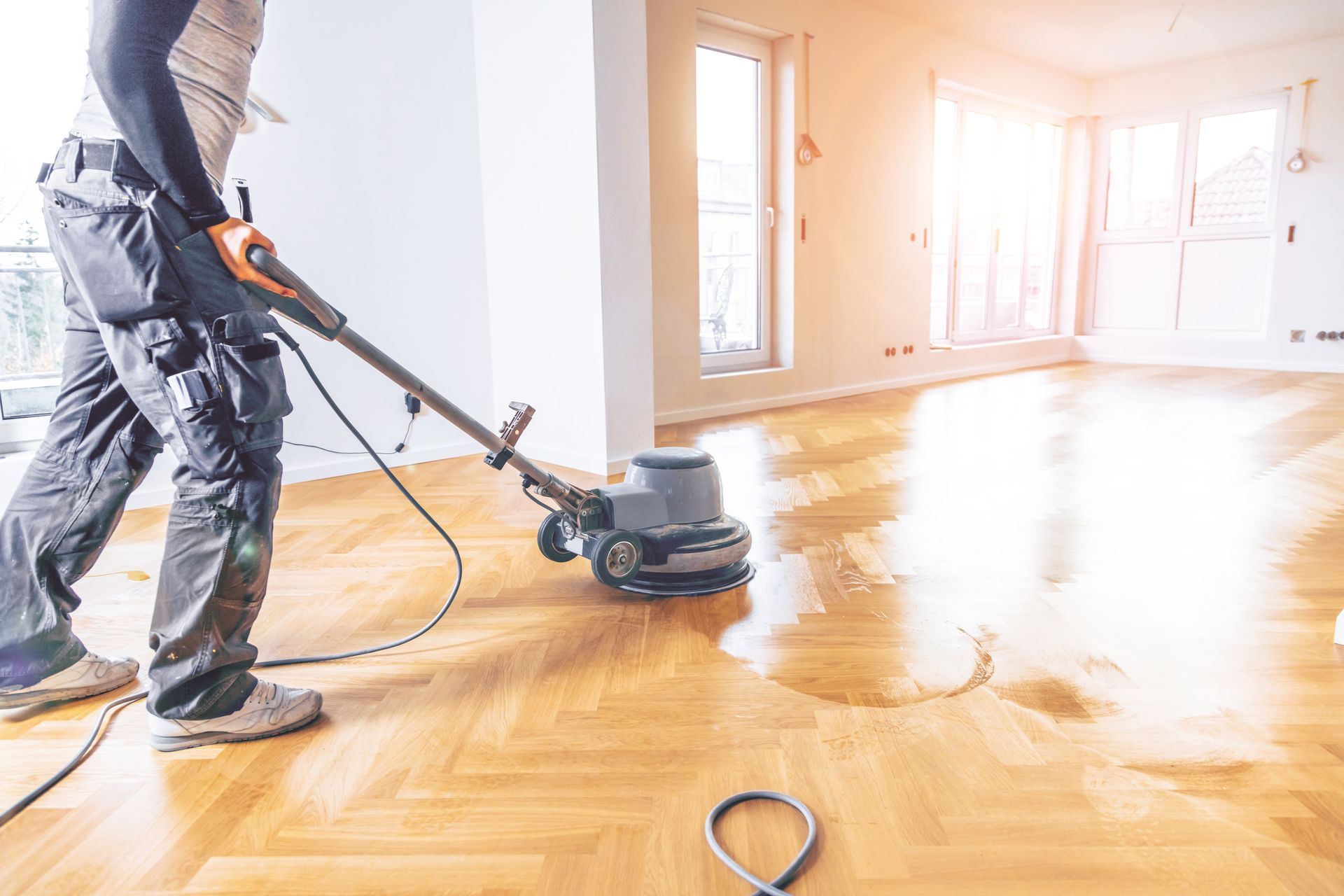 Person buffing a sunlit hardwood floor with a floor polisher in an empty room