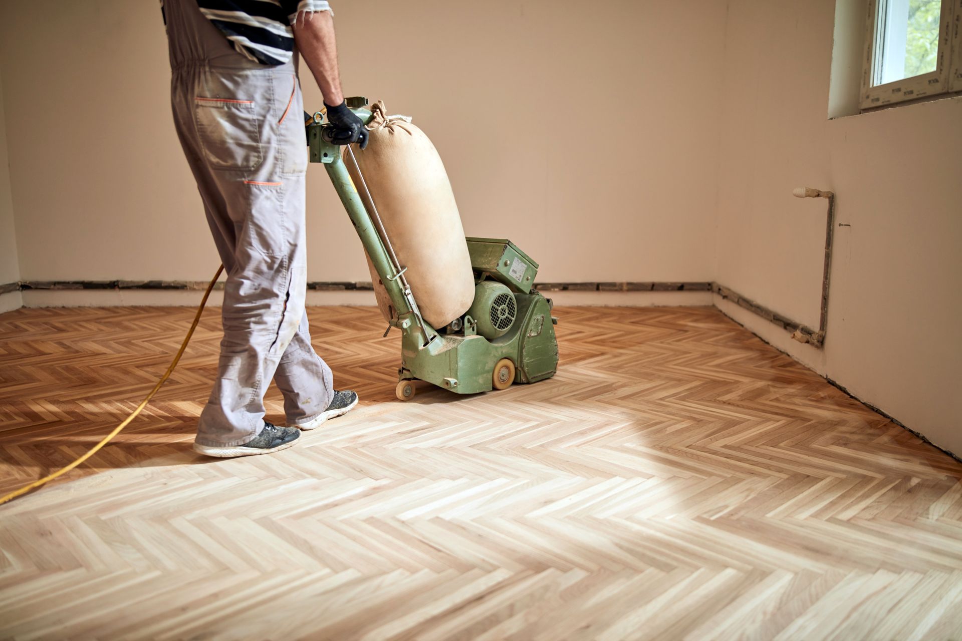 A man is refinishing a parquet wood floor using a heavy-duty drum sanding machine.