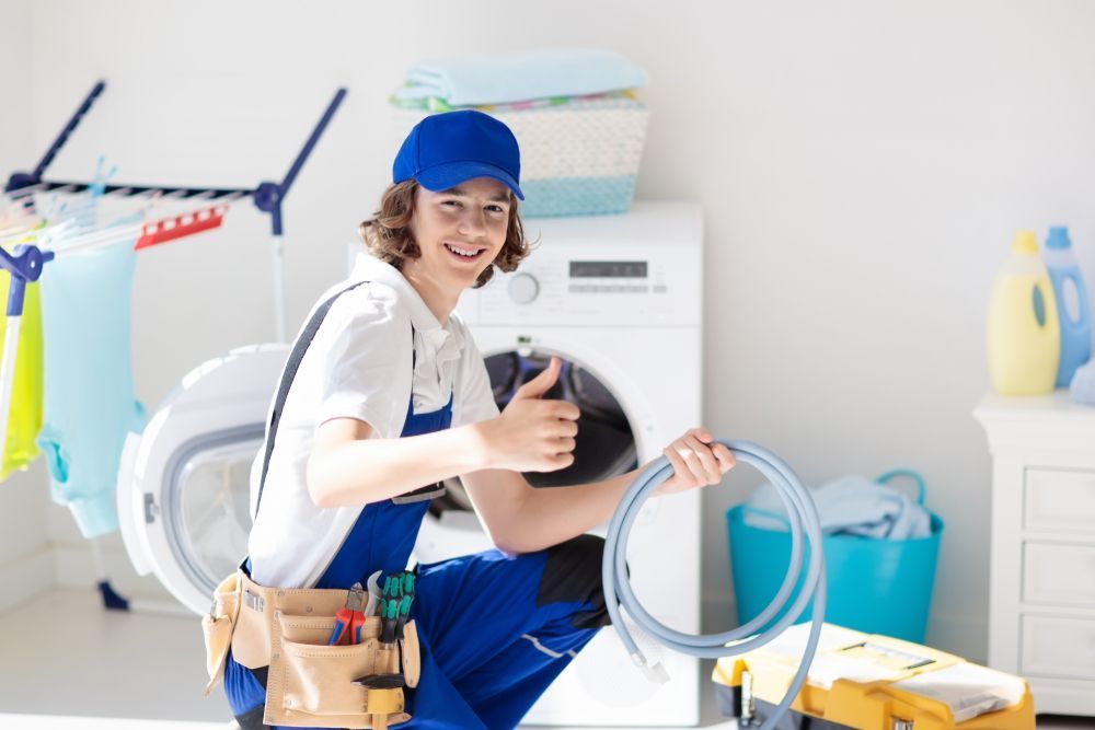 Repair technician in blue overalls giving a thumbs up in a laundry room, holding a hose.