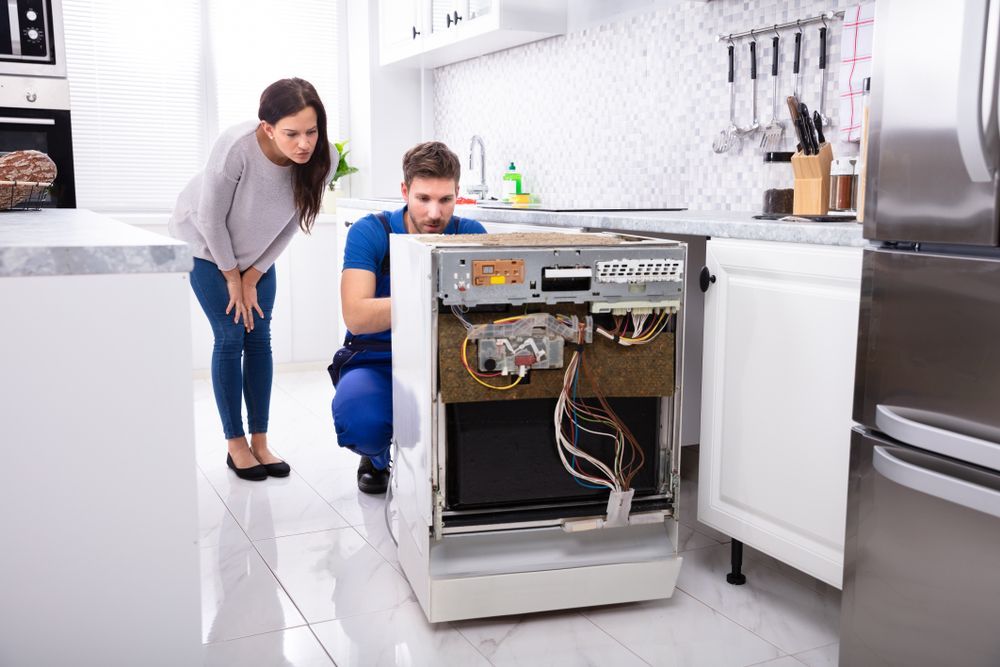 A repairman fixes a dishwasher in a kitchen, a woman watches, concerned.