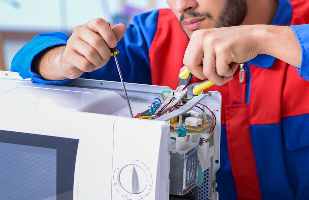 Technician repairing a microwave using a screwdriver and pliers, wearing blue and red overalls.