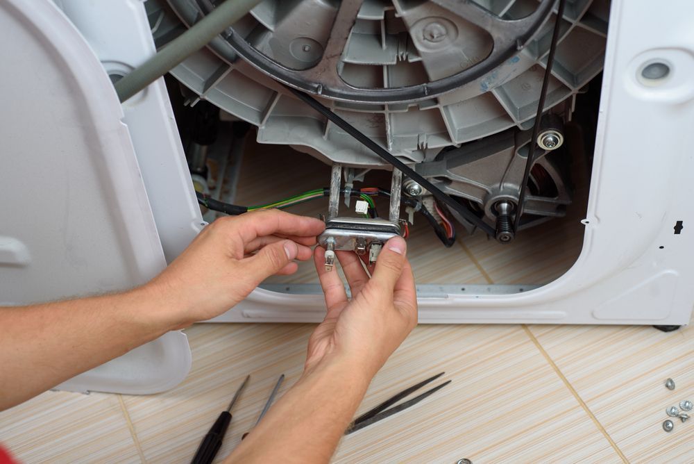 Hands repairing a washing machine, holding a metal part. Tools and interior machine details visible.