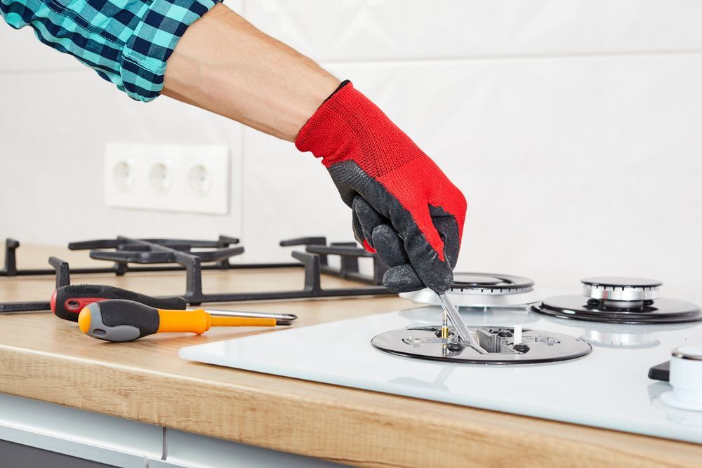 Person in red glove repairs a white stovetop with tools on a wooden counter.