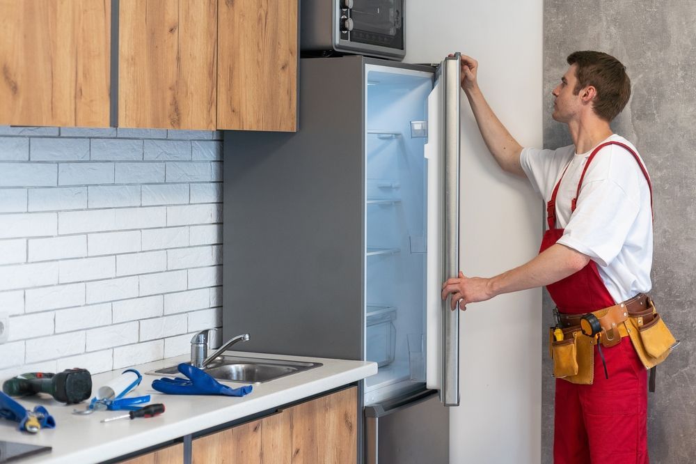 Man in red overalls opens refrigerator in a kitchen with tools on the counter.