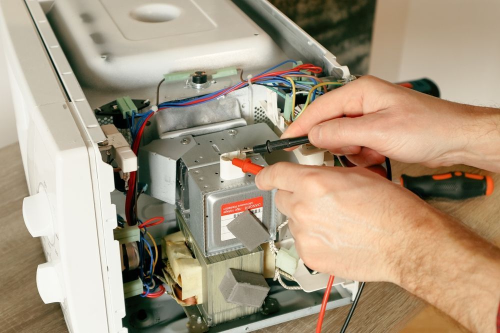 Person testing a microwave's internal components with a multimeter, revealing wires and electronics.