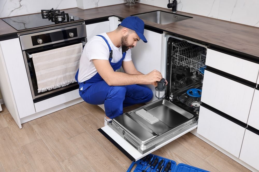 A technician in blue overalls repairs a dishwasher in a kitchen.