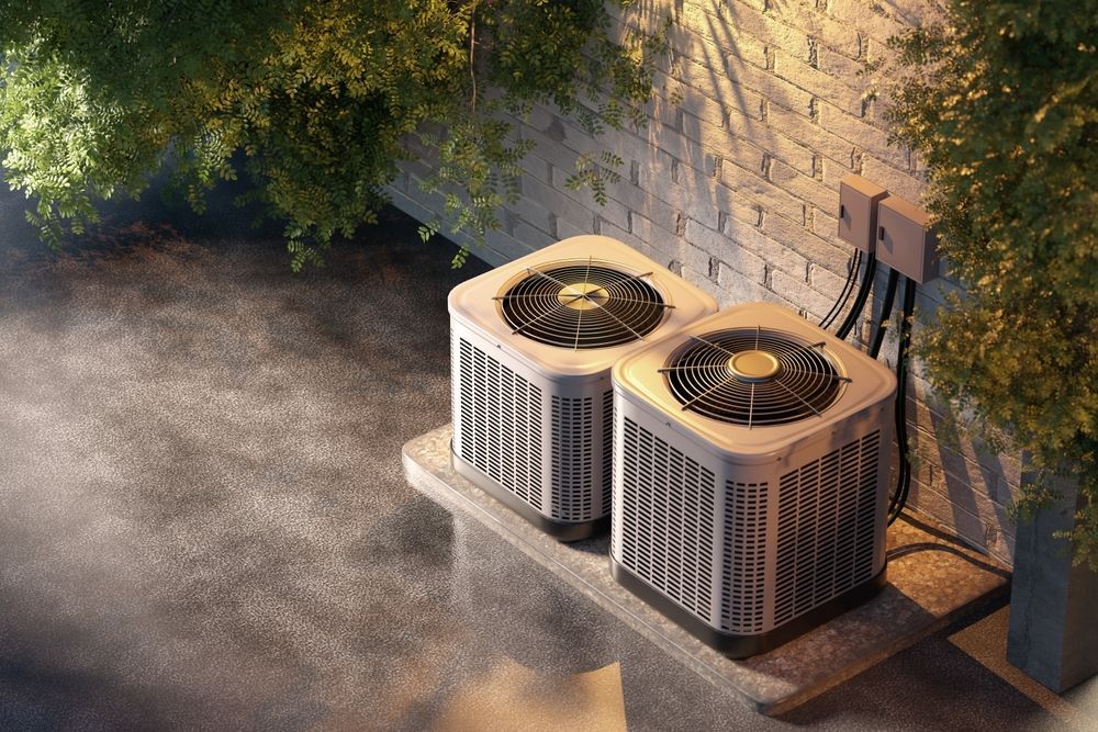 Two air conditioning units on a concrete pad near a brick wall, with greenery around.