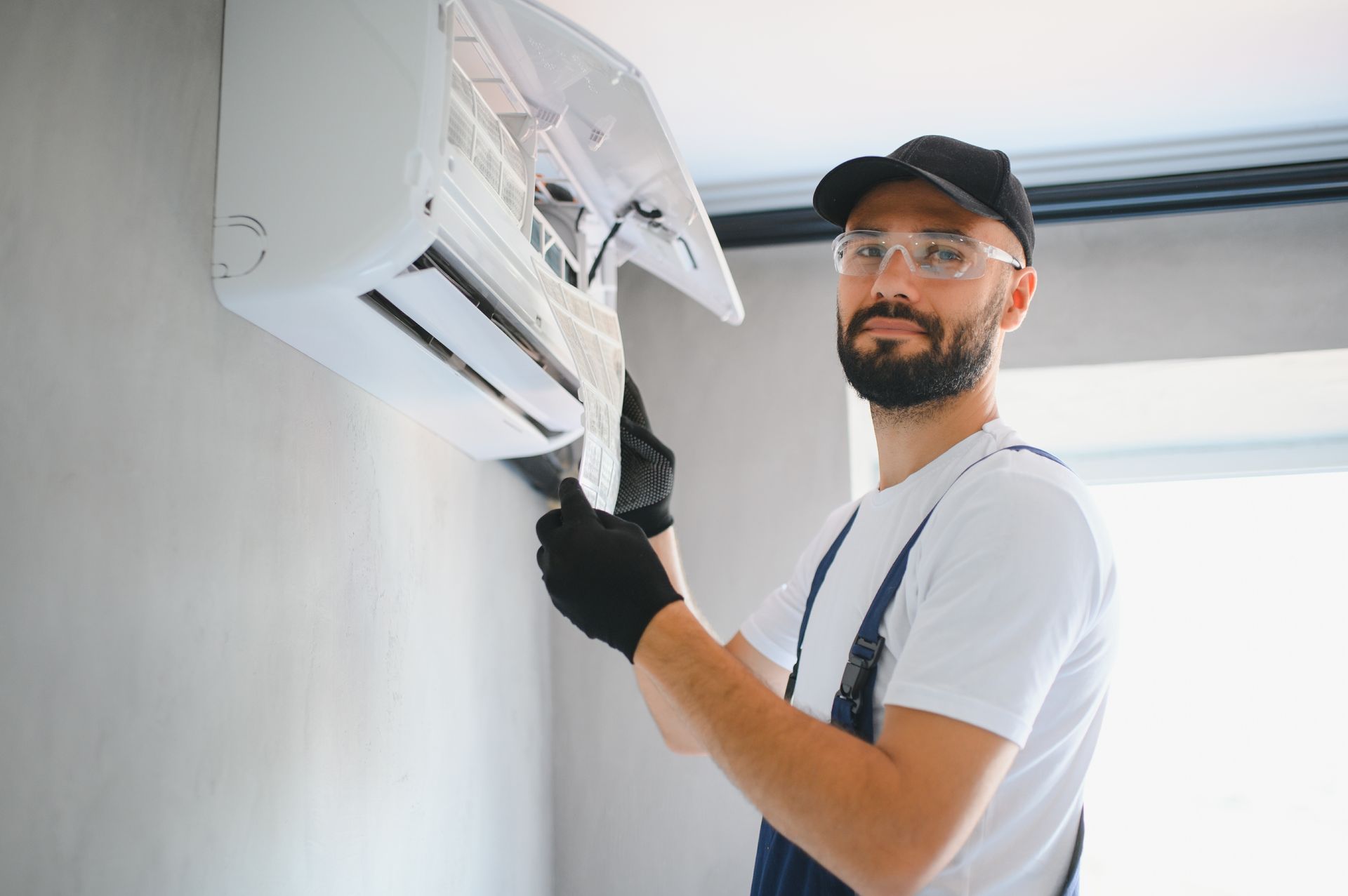 Man in overalls fixing an air conditioner unit on a gray wall.