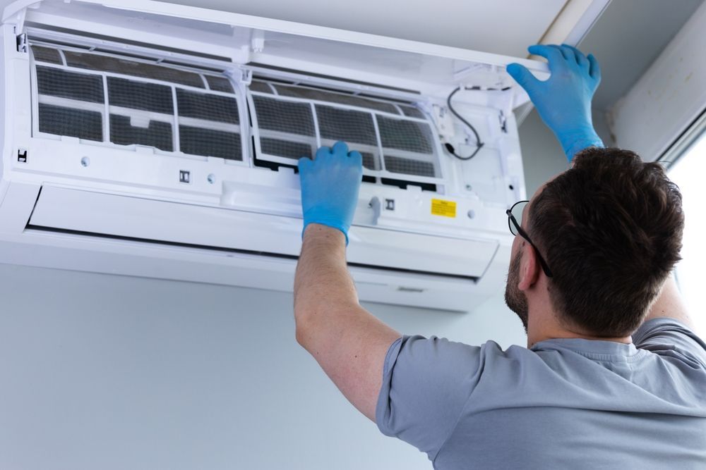 Person in blue gloves cleaning the air filter of a wall-mounted AC unit.