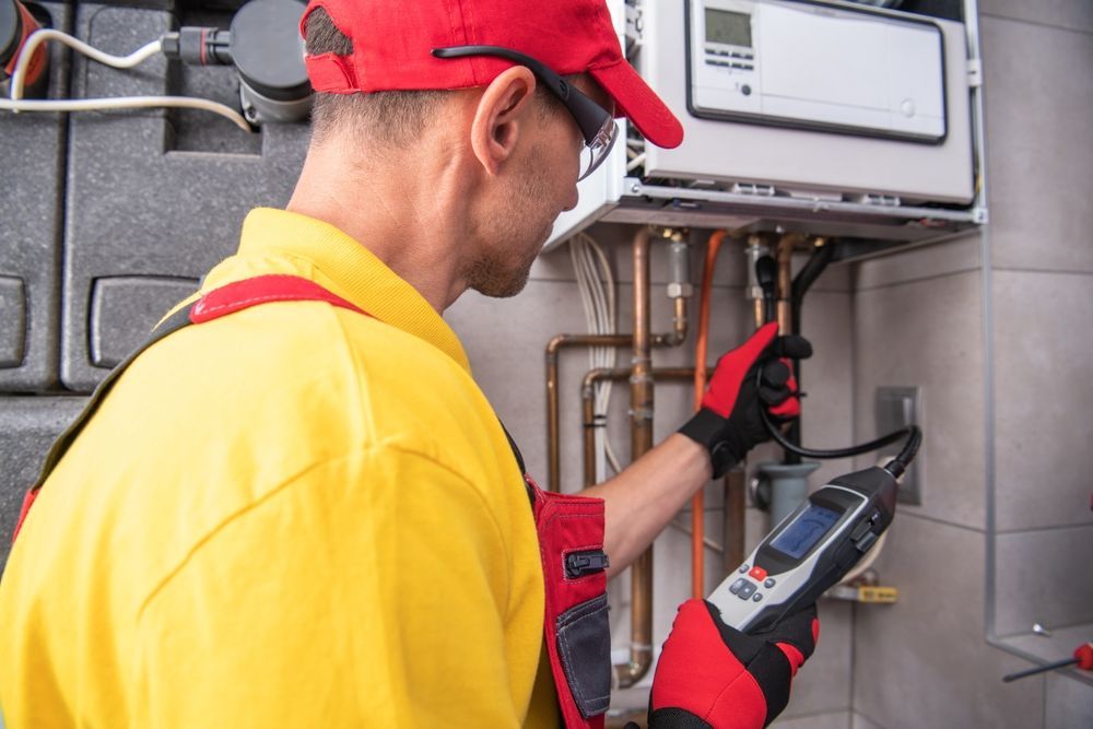 A person in yellow and red overalls uses a device to inspect a wall-mounted boiler with copper pipes.