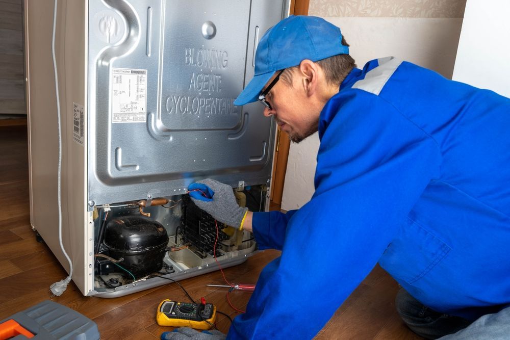 A person in blue overalls repairs a refrigerator's compressor; they are wearing a cap and gloves.