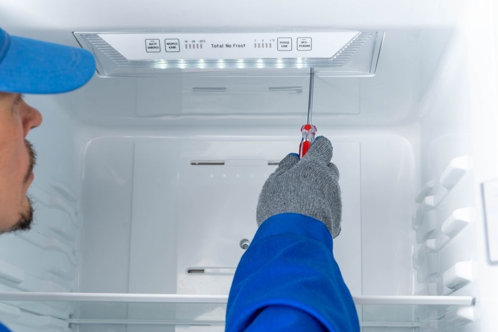 Person in blue uniform fixing refrigerator light with a screwdriver.