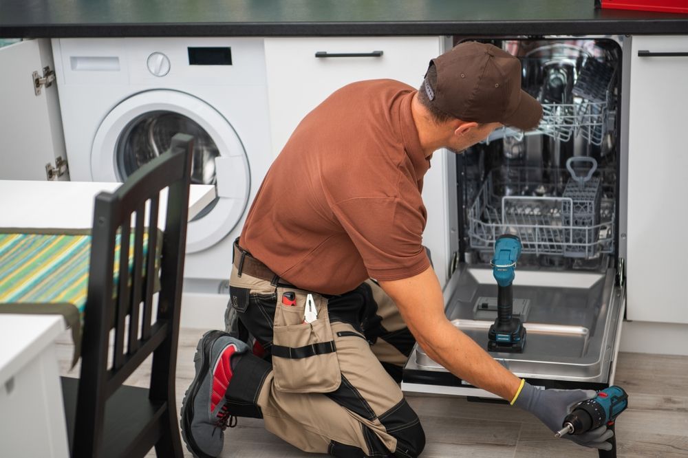 A person repairing a dishwasher in a kitchen. They wear a brown shirt, cap, and work gloves. A washing machine is visible.