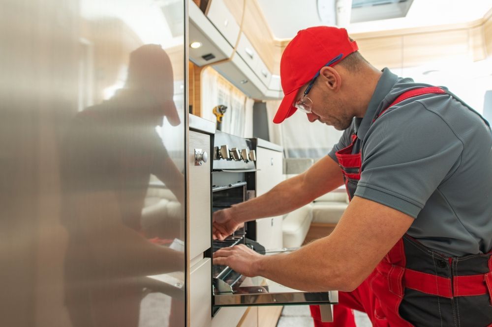 Technician in red overalls and hat repairs oven inside a kitchen.
