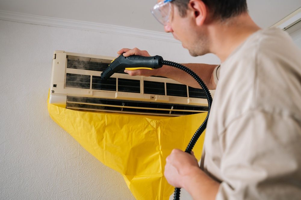 A person cleaning an AC unit with a steam cleaner, using a yellow cover to catch debris.