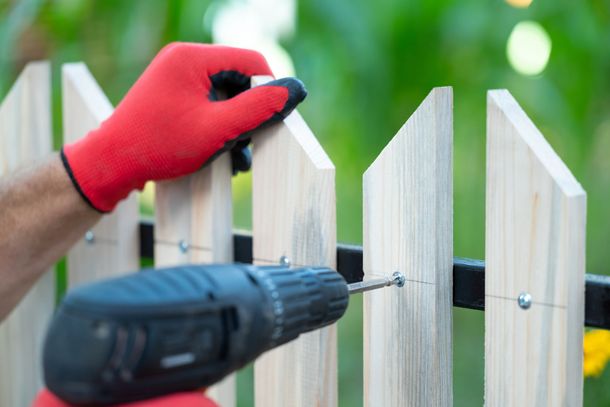 A hand in a red work glove holds a wooden picket while a power drill fastens it to a black fence rail.