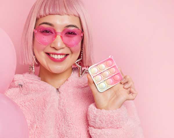 A woman with pink hair and sunglasses is holding a pink pop it toy.