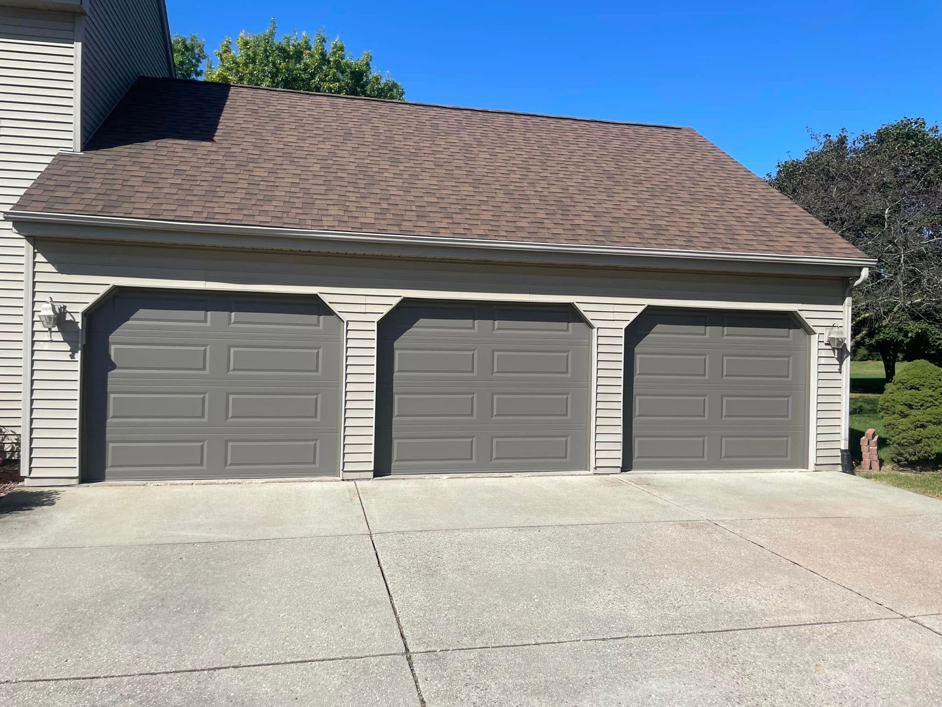 Three-car beige garage with gray doors and brown shingled roof, viewed from the driveway.