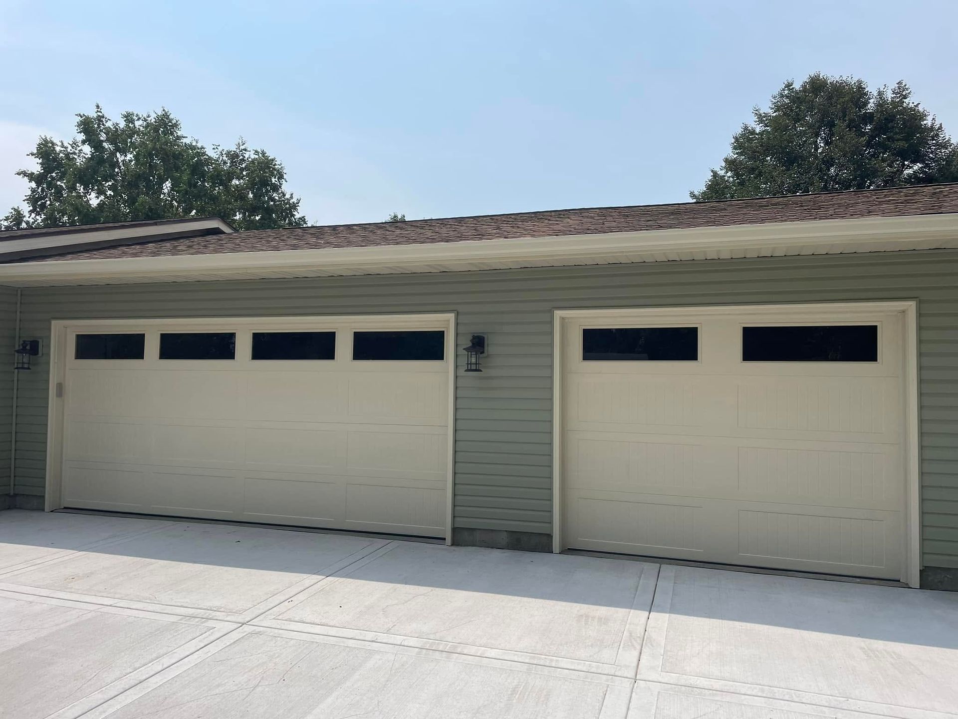 Two cream-colored garage doors on a light green house with a concrete driveway and trees above.