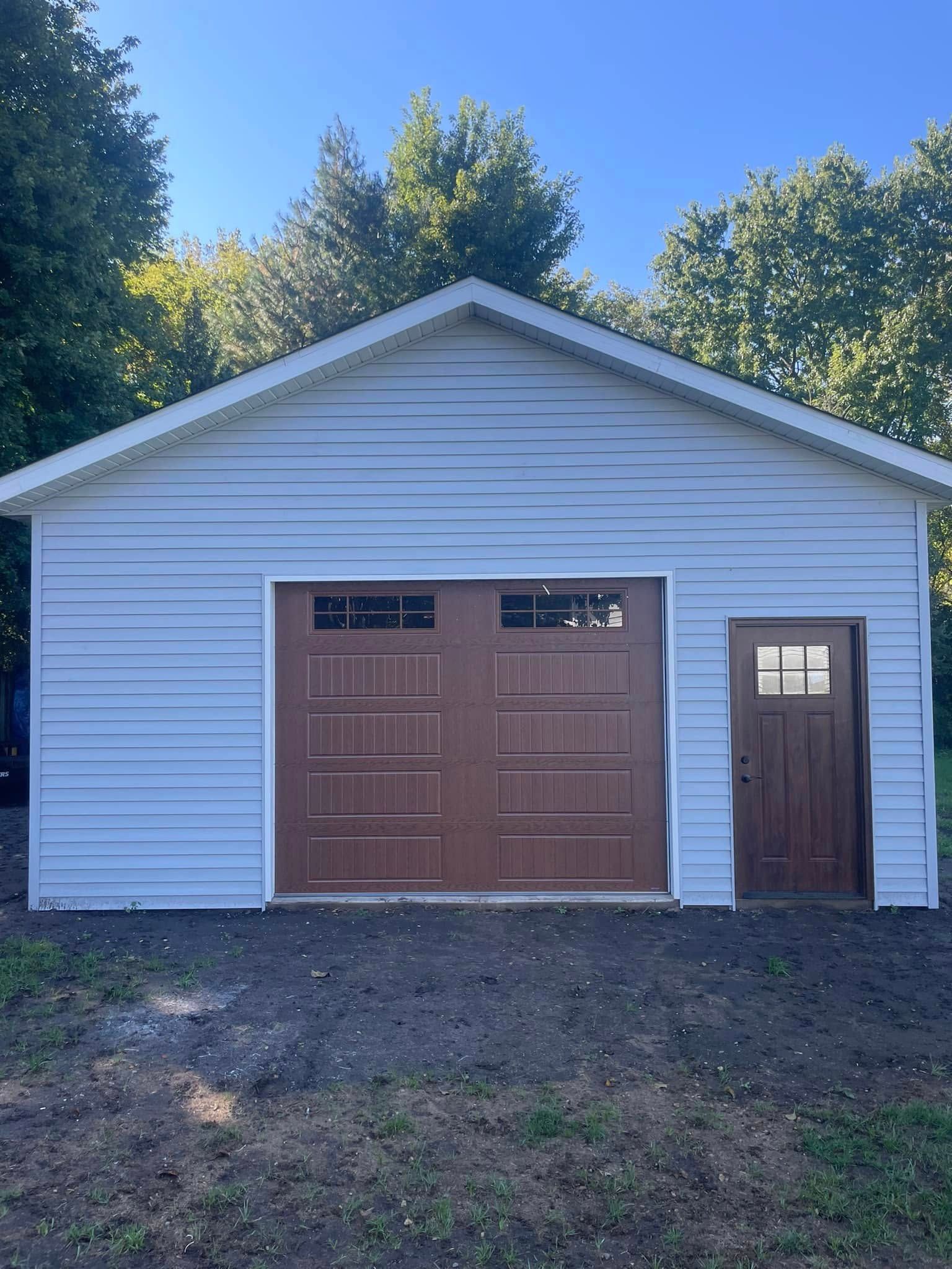 White garage with brown double doors and a side entry door, set on a grassy driveway under blue sky.