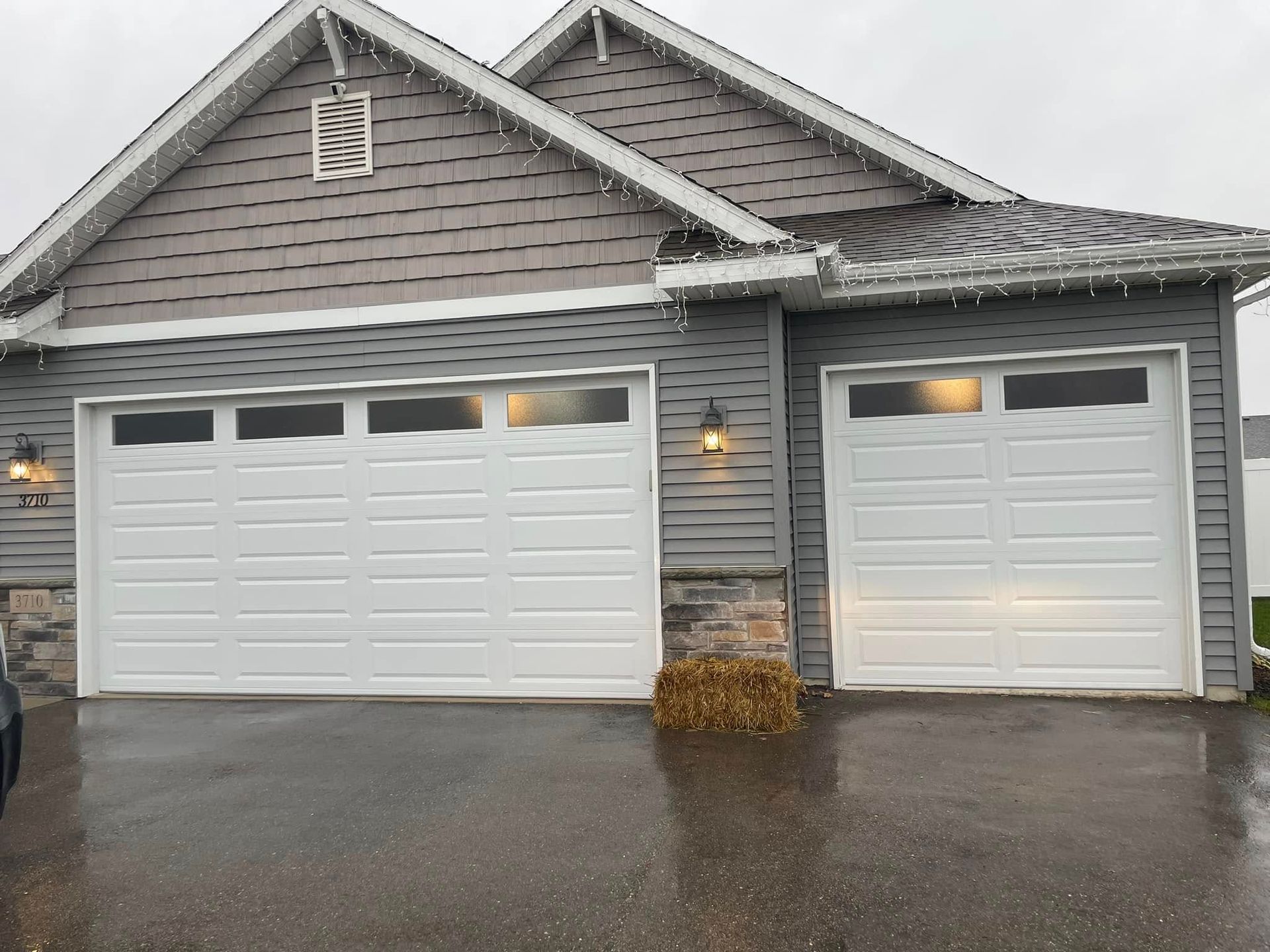 Two-car garage with white doors on a gray house, seen from a wet driveway in light rain.