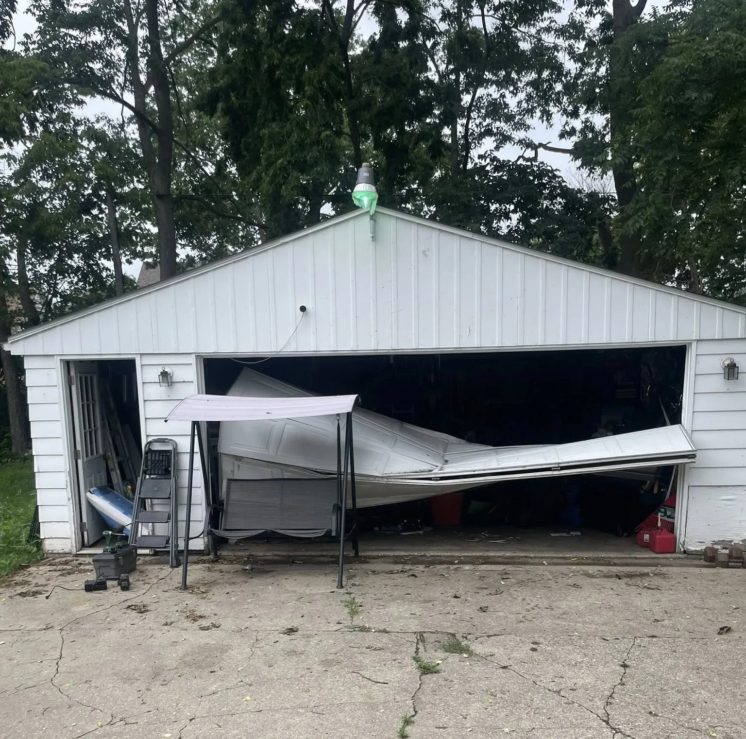 White garage with a collapsed front opening and tarp-covered items in the driveway, trees behind.