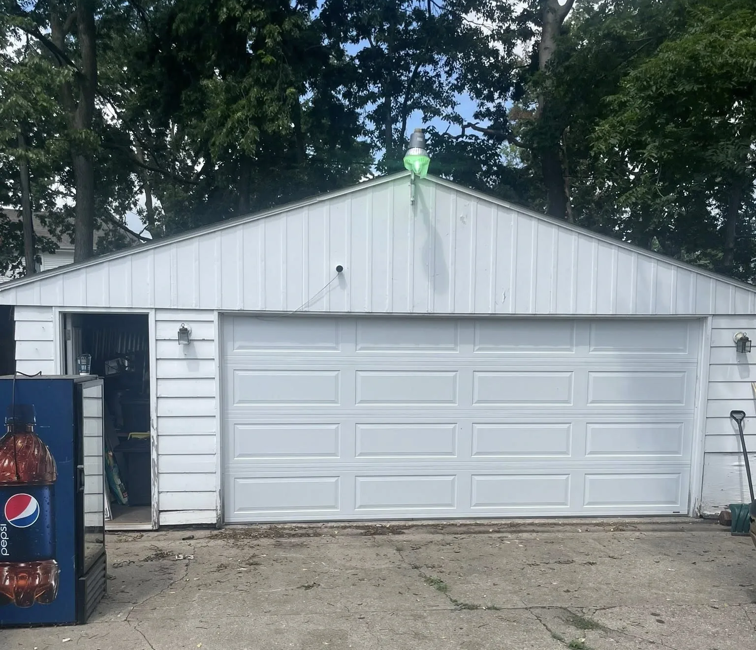 White garage with closed double door and a small green light on top, framed by trees and a driveway.